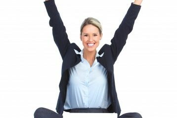Woman sitting on the ground in front of her laptop with her arms up in celebration while she smiles at the camera