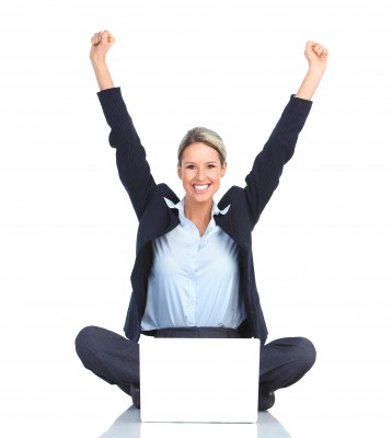Woman sitting on the ground in front of her laptop with her arms up in celebration while she smiles at the camera