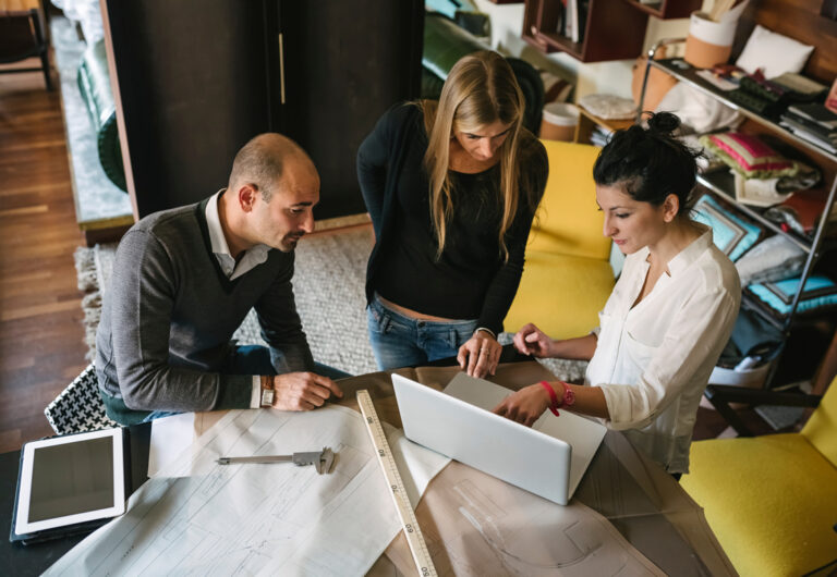 3 designers looking at a laptop at a drawing table with blueprints in front of them