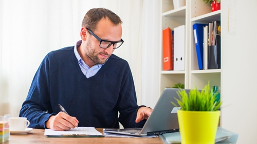 man writing on a clipboard as he scrolls on his laptop