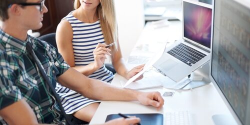 Coworkers sitting at a desk in front of their computers discussing